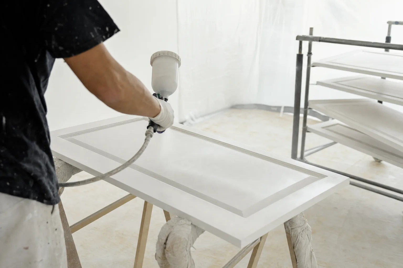 Painter spraying a smooth white finish on a kitchen cabinet door on sawhorses