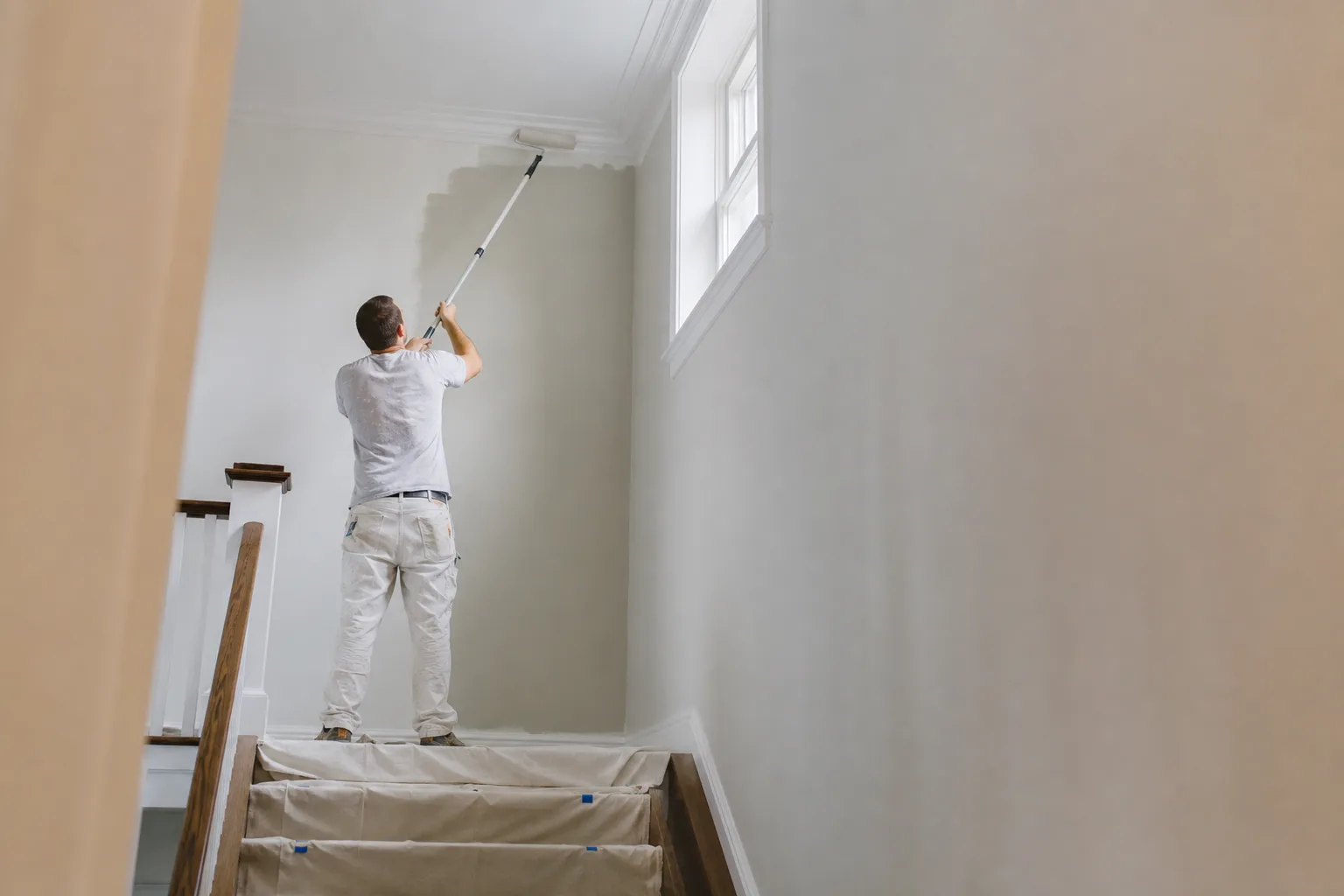 Painter using an extension pole and a stair platform to roll the high wall of a tall interior stairwell