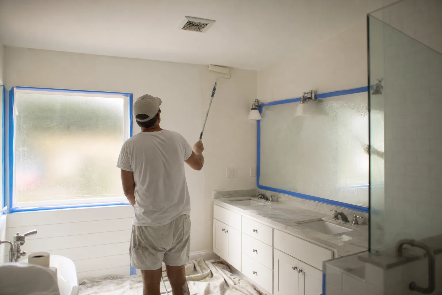 Painter rolling mildew-resistant satin paint onto a humid Gulf Coast bathroom wall and ceiling near an exhaust vent