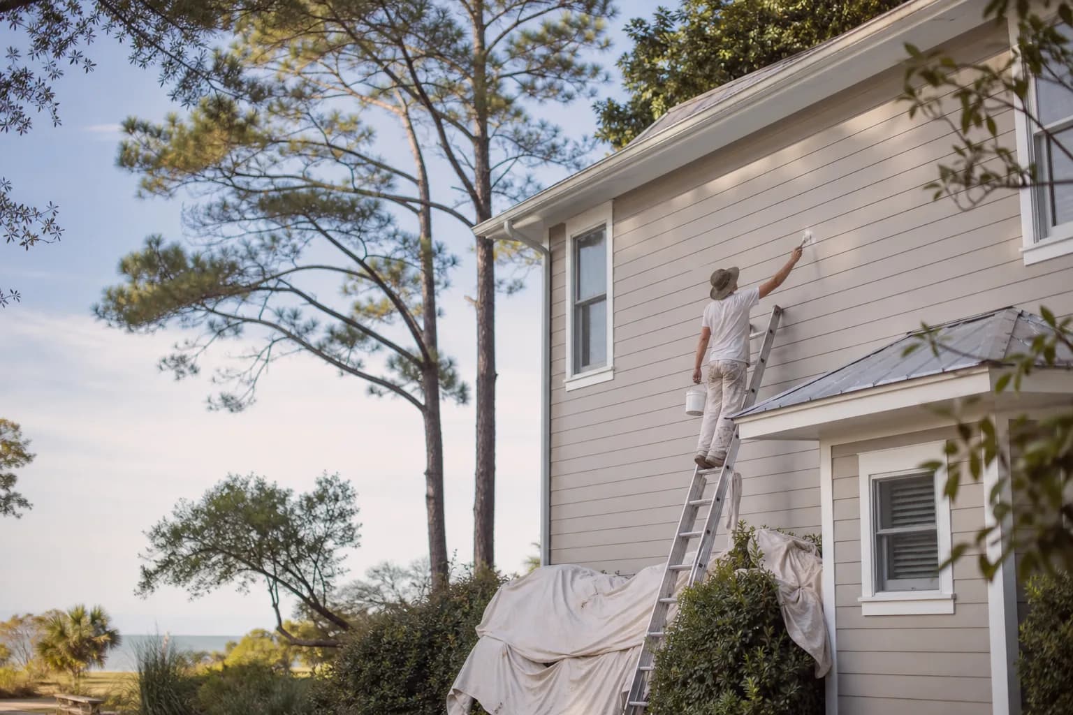 Painter prepping and coating the trim of a two-story Baldwin County home, illustrating what drives exterior house painting cost